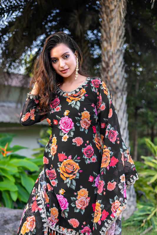 Elegant woman in black floral salwar suit with vibrant roses, showcasing style against a lush backdrop.