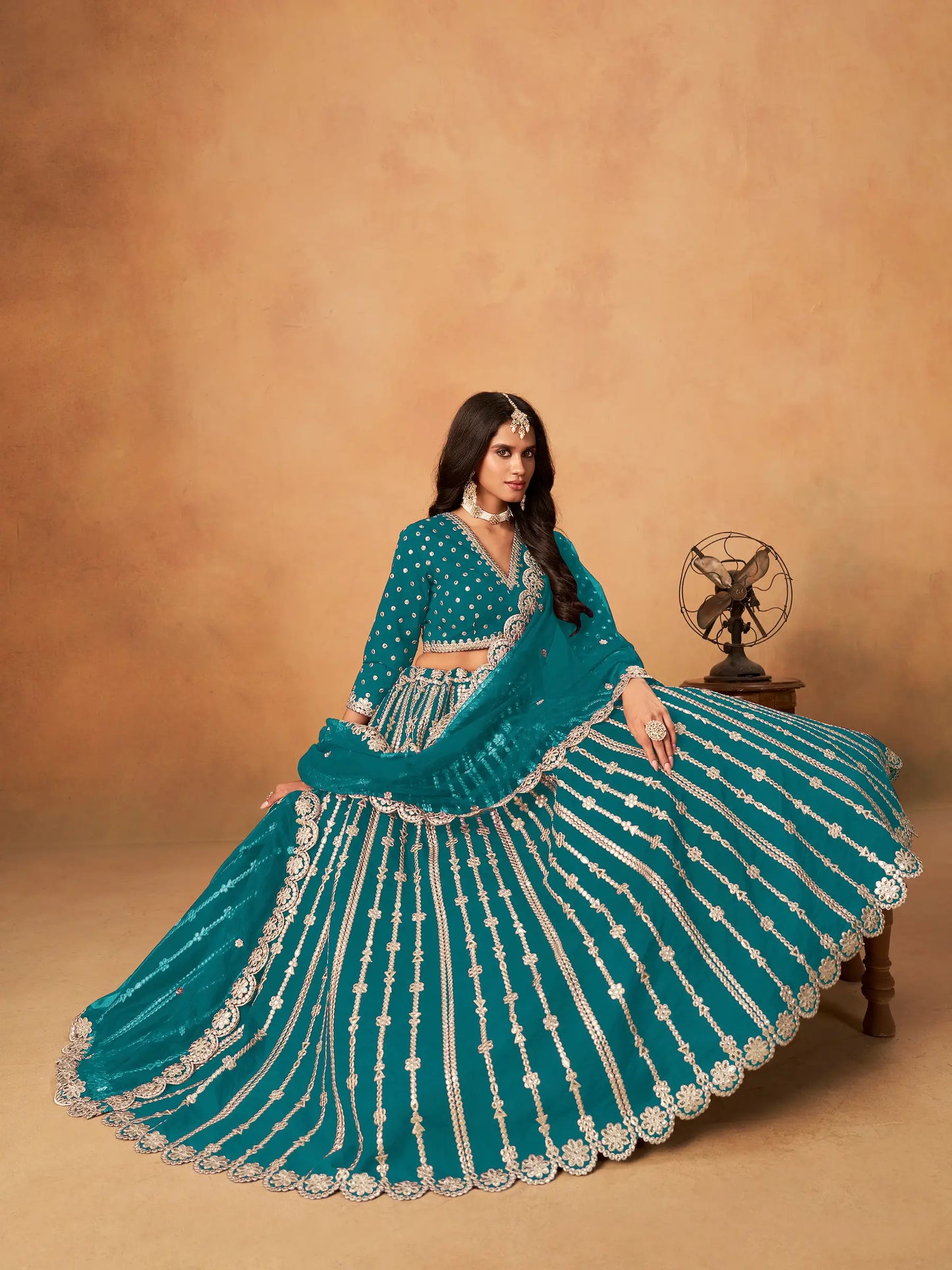 Model sitting wearing teal blue art silk lehenga choli with paper mirror and embroidery work